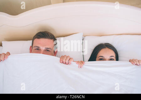 A picture of man and woman hiding faces under white blanket. They are lying together in one bed. Peple are looking up straight forward Stock Photo