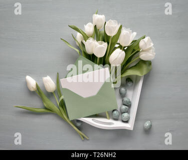 Blank card in paper envelope with the bunch of white tulips on a decorative tray with Easter eggs, toned image, text space on the card. Stock Photo