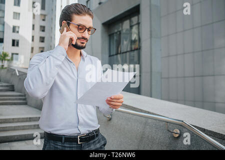 Bearded businessman in sunglasses stands on steps and hold piece of paper. He looks at document. Guy studies it. e talkes on phone at the same time Stock Photo