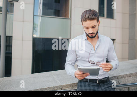 Serious and concentrated young bsinessman leans to railing. He holds talet in hands and looks at it. Also guy has sunglasses in left hand Stock Photo