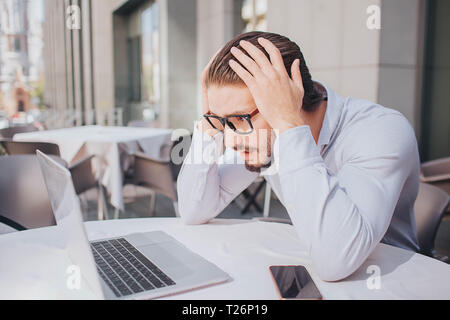 Busy and uhappy young man looks at screen of laptop closer. He holds hands on head. Guy is amazed. There are his phone at table Stock Photo