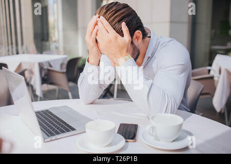 Unhappy and sad young man sits at table in restaurant. He closed his face with hands. Guy is emotional. There are two cup and phone with laptop at tab Stock Photo