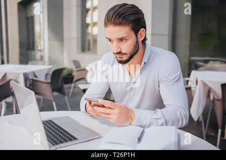 Concentrated and calm young man sits attable and looks at laptop's screen. He holds phone in hands. There are opened notebook with pen on table. He wo Stock Photo