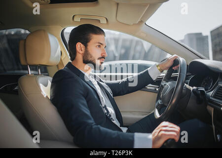Handsome and confident young man sits in car and looks straight forward. He drives car. It looks luxury inside. He holds one hand on steering wheel an Stock Photo
