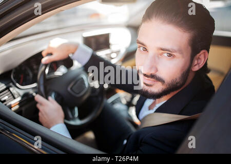 Satisfied and happy man sitting in car and posing. He looks at camera and smiles a bit. Bearded guy holds hands on steering wheel Stock Photo