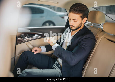 Busy young man sitting in luxury car and looking at watches. He is late. Guy wears suit. He is serious and concentrated Stock Photo