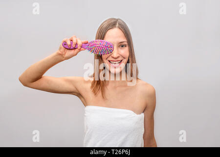 Nice and positive model stands and poses on camera. She smiles. Woman coveres one eye with pink brush. Her body coveres with white towel. Isolated on  Stock Photo