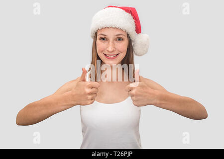 Positive young woman looks on camera and shows her big thumbs up. She wears white t-shirt and red Christmas hat. Model smiles. Isolated on grey backgr Stock Photo