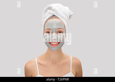 Positive and happy young woman looks on camera and smiles. She has mask on her face. Hair is covered with white towel. Isolated on grey background Stock Photo