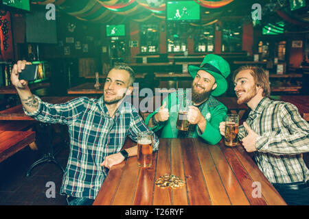 Three young men sit at table in pub and take selfie. They pose on camera and smile. Guy in middle hold phone Stock Photo