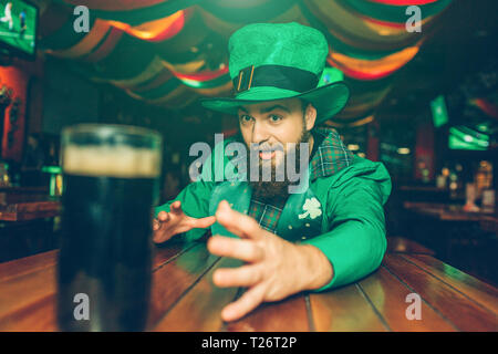 Afraid bearded guy in green suit sit at table in pub. He reach mug of dark beer with hand. Young man wear St. Patrick's suit Stock Photo