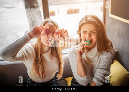 Funny young women cover eyes with macarons. They pose on camera. Brunette bites piece of macarone. They have fun Stock Photo