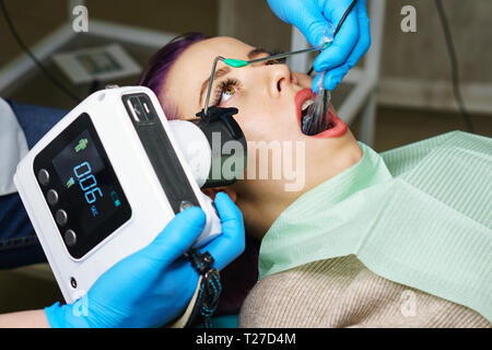 Dentist checks thickness of tooth enamel using portable x-ray. Young girl with open mouth. White teeth. Dentist hands closeup. Dental clinic. Stock Photo