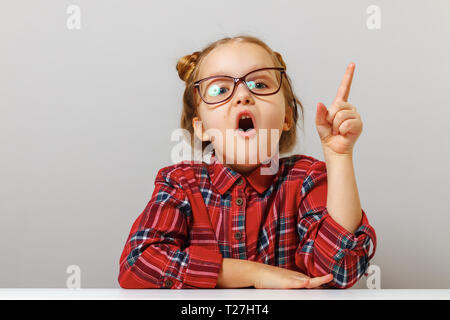 Funny little girl with glasses points finger up. Back to school. Education concept. Gray background. Stock Photo