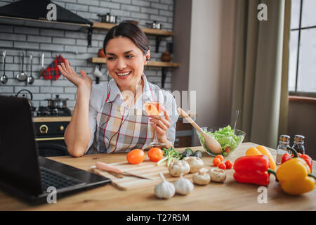 Nice positive woman sit at table in kichen. She talk through headphones and wave to laptop screen. Woman happy. Vegetables lying on table Stock Photo