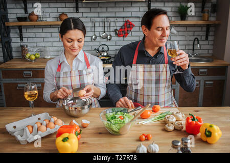 Nice couple sit together at table in room. Woman break eggs above bowl. Man drink white wine from glass. They spend time together Stock Photo