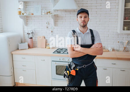 Plumber in kitchen. Professional look on camera and pose. Hands crossed. Tools on belt aside Stock Photo