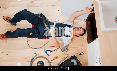 Concentrated busy plumber fixing on in kitchen under sink. He lying on floor. Guy hold wrench Stock Photo