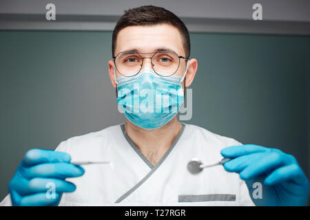 Serious young male dentist wear uniform and mask. He hold dental tools in hands with latex gloves. Guy look on camera. Isolated on green background Stock Photo