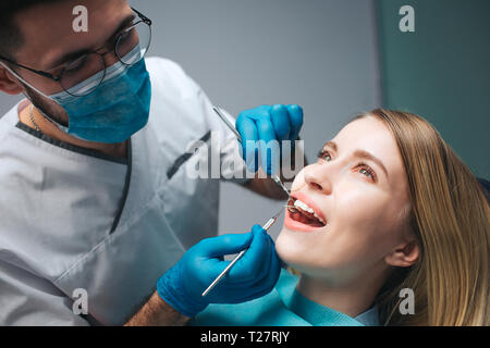 Close up of dentist checking client's teeth with special tools. Young woman sit in chair in room and keep mouth opened. She look up. Stock Photo