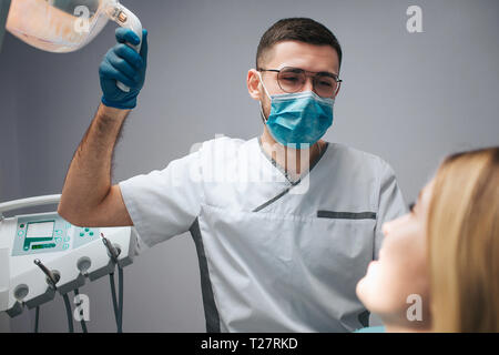 Young dentist in mask sit at client and hold lamp. Young woman look at doctor and smile. They are in dentistry room.
