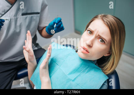 Young woman look afraid. She sit in chair in dentistry. Woman look on camera and push away doctor. He hold dentist's tool.