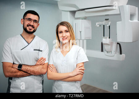 Two dentists stand in x-ray room and pose to camera. They smile and hold hands cossed. Professionals on picture. Stock Photo