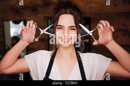 Portrait of smiling beautiful woman hairdresser with black apron looking at camera while holding professional scissors on salon background. Beauty and Stock Photo