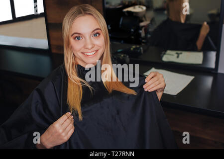 Smiling blonde young woman with black cape sitting in hairdressers chair in front of mirror while waiting for new haitcut in salon. Beauty and people Stock Photo