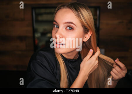Portrait of blonde young woman with black salon cape looking at camera while waiting for a hairdresser on salon background Stock Photo