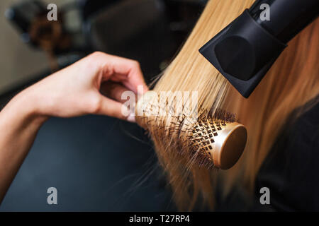 Close-up of hairdressers hand drying blond hair with hair dryer and round brush. Doing hairstyle in beauty salon Stock Photo