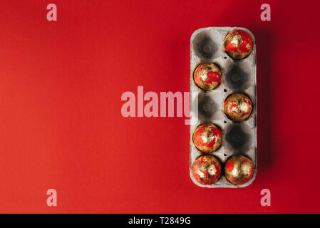 Six golden colored and decorated with sparkles Easter eggs in cardboard egg-box, Easter egg hunt celebration, colorful still life in paper box on red  Stock Photo