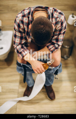 Man with bottle of beer sitting on toilet with his pants down, top view, after house party. Drunk male person with headache
