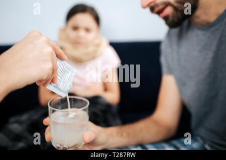 Cut view of doctor's hand pouring powder medicine into glass of water. Young man hld it. Small sick girl sit on sofa and look at process Stock Photo