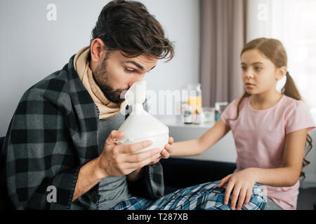 Sick young man doing inhalation procedures. He hold special equipment fo this. Worried small girl sit besides him in one room Stock Photo