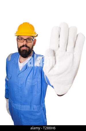 Fun high angle of bearded workman with yellow helmet, blue uniform and in glasses making stop gesture with his hand in gloves. Isolated over white bac Stock Photo