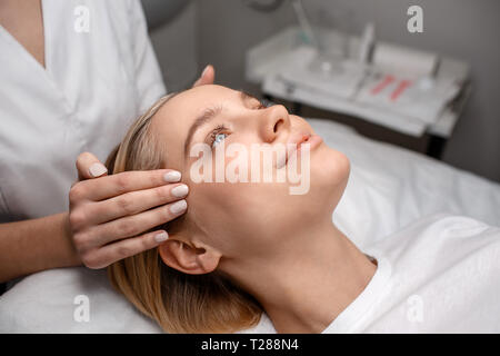 Nice young woman lying on couch in room. Beautician hands doing head massage. Model smile. Stock Photo