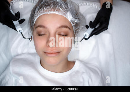 Relaxed peaceful young woman lying on couch with closed eyes. She enjoy. Beautician's hands touching face with beauty equipment. Stock Photo