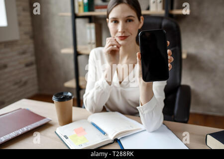 Attractive young businesswoman show phone on camera. She sit at table in room. Opened notebook and cup of coffee. Stock Photo