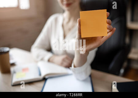 Nice young businesswoman hold yellow stickers in hand. She show them to camera. Model sit in room. Daylight. Stock Photo