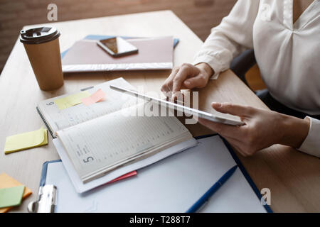 Cut view of woman's hands holding tablet in hands on table. Notebooks lying there with cup of coffee and phone. Busy work. Stock Photo