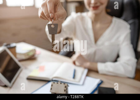 Young businesswoman sit at table in room. She hold key and smile. Busy woman work. Notebook and tablet on table. Stock Photo