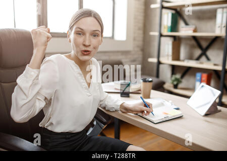 Cheerful nice young woman sit at table in room. She happy. Model look on camera and hold fist. Stock Photo