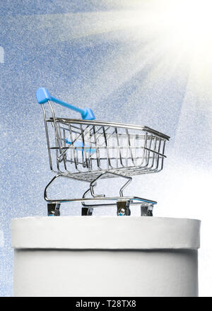 Shopping trolley on a round white gift box on a winter blue shiny background with bright rays. The best buyer or the best purchase. Stock Photo