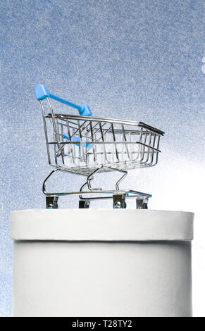 Shopping trolley on a round white gift box on a winter blue shiny background. The best buyer or the best purchase. Stock Photo