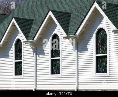 Oblique exterior view  of dark green roof and bright white wall of church, with three triangular roof gables above three stained glass windows Stock Photo