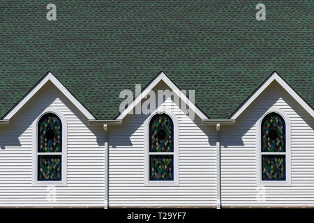 Exterior view of dark green roof and bright white wall of church, with three triangular roof gables above three stained glass windows Stock Photo