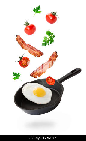 Fried egg, bacon and cherry tomatoes flying into iron skillet isolated on white background. Stock Photo
