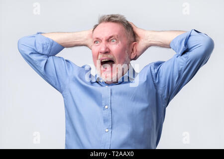 Senior man in blue shirt with angry grimace on his face,with mouth opened in shout, ready to argue. Studio shoot. Negative human emotion. Stock Photo