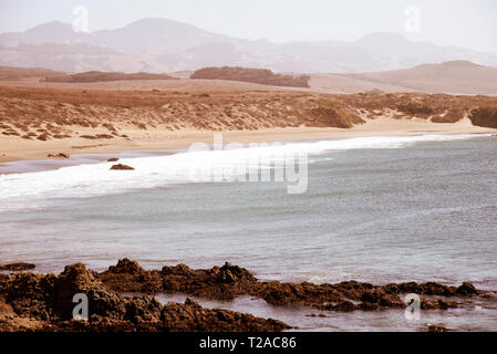 Overlooking rocky shoreline with ocean waves breaking onto sandy beach and hazy mountains beyond under bright hazy skies. Stock Photo